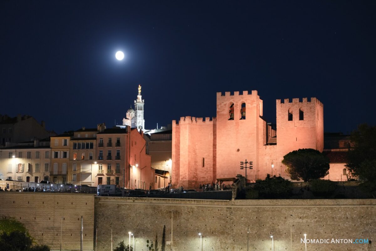 Moon above illuminated stone towers, with Notre-Dame de la Garde lit in the background.