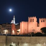 Moon above illuminated stone towers, with Notre-Dame de la Garde lit in the background.