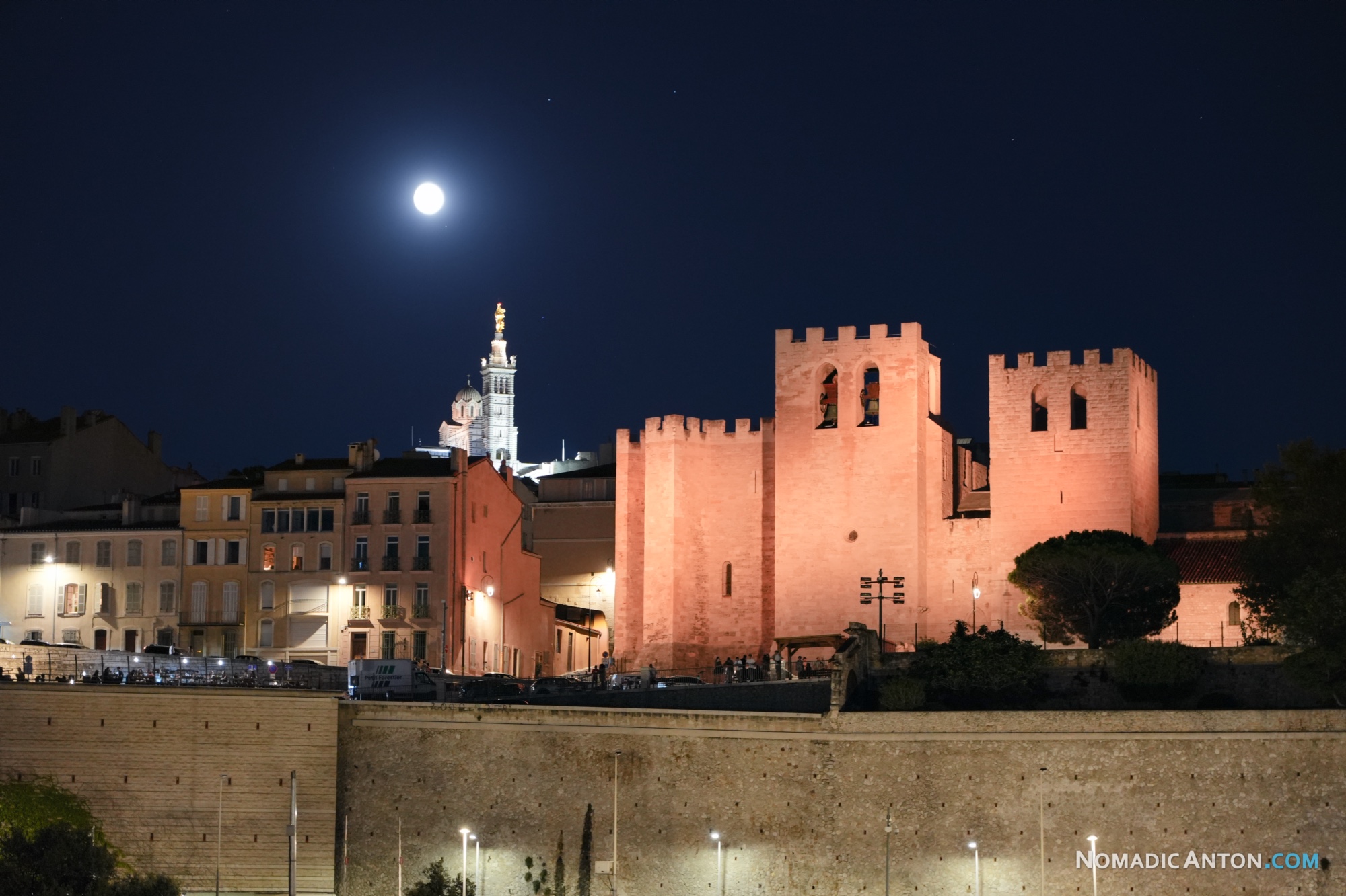 Moon above illuminated stone towers, with Notre-Dame de la Garde lit in the background.