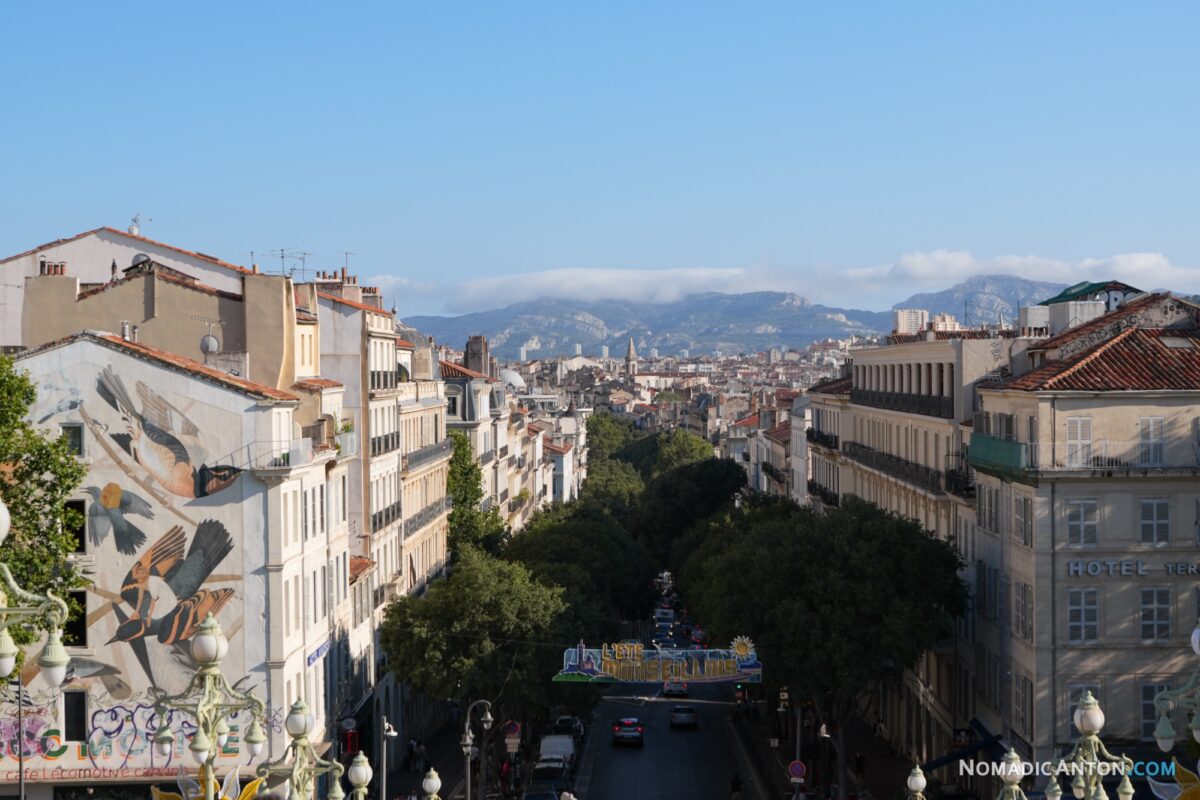 View down a Marseille street past rooftops and a large wall mural toward distant hills.