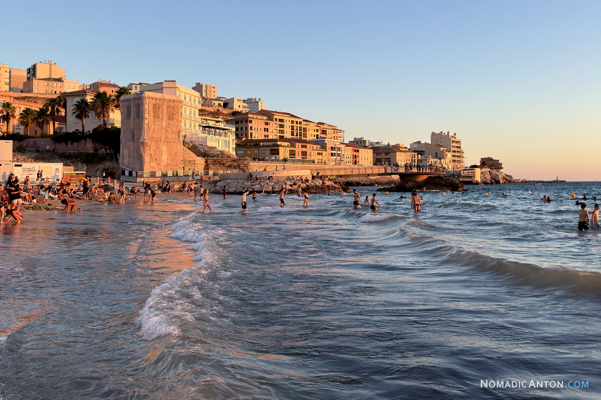 People swimming and wading at Plage des Catalans as waves reach the sand beneath sunlit waterfront buildings