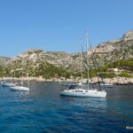 Sailboats on blue water beside the rocky slopes of a calanque.
