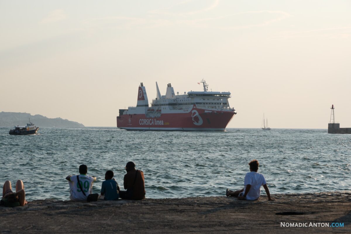 People sitting beside the sea as a red ferry passes in the evening light.