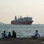 People sitting beside the sea as a red ferry passes in the evening light.