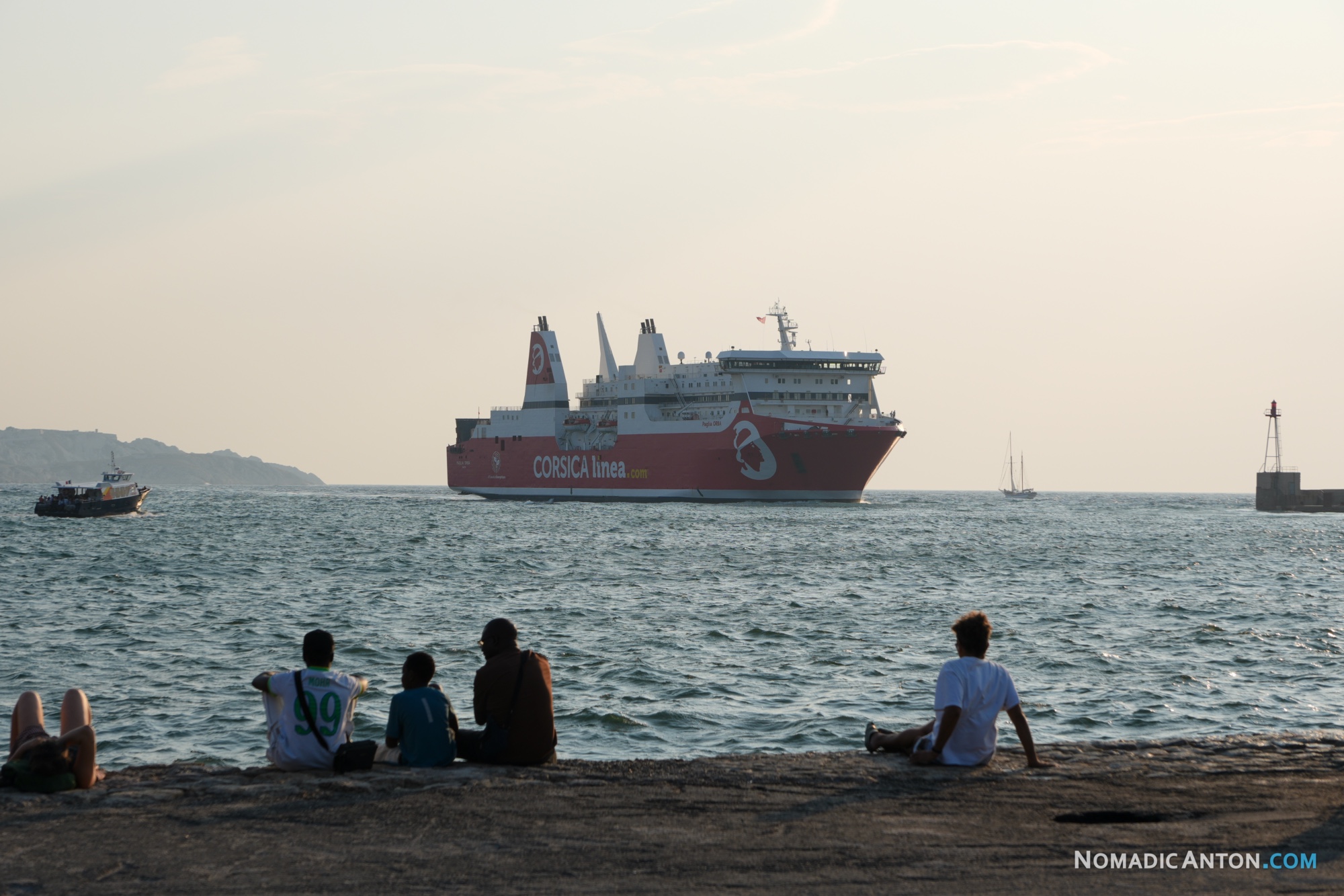 People sitting beside the sea as a red ferry passes in the evening light.